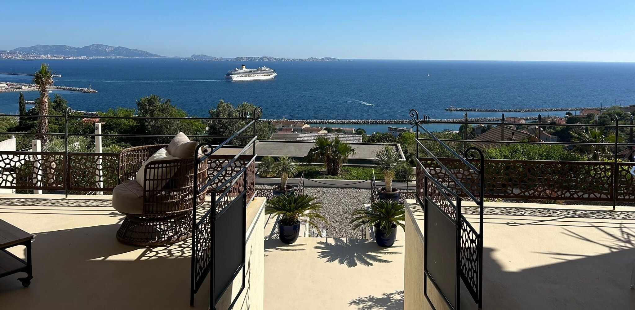 La terrasse couverte de la villa : un fauteuil en rotin, la rambarde ajourée, et toute la rade de Marseille avec un paquebot au large.